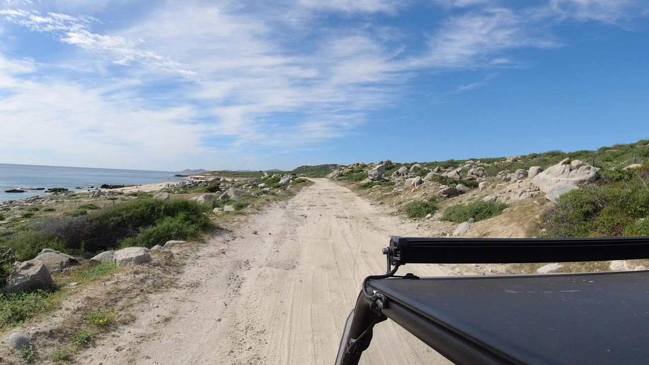 Black vehicle hood on a sandy coastal dirt road beside rocks and sea.