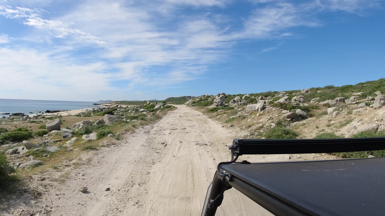 Vehicle foreground on a dusty track with boulders and coastline under blue sky.