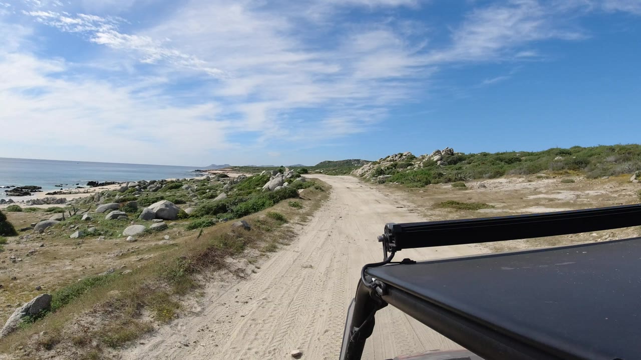 Los Cabos, Mexico dirt road curves ahead between coastal rocks and scrub, with blue ocean on the left, a black vehicle roof edge in the lower right, and no visible people.