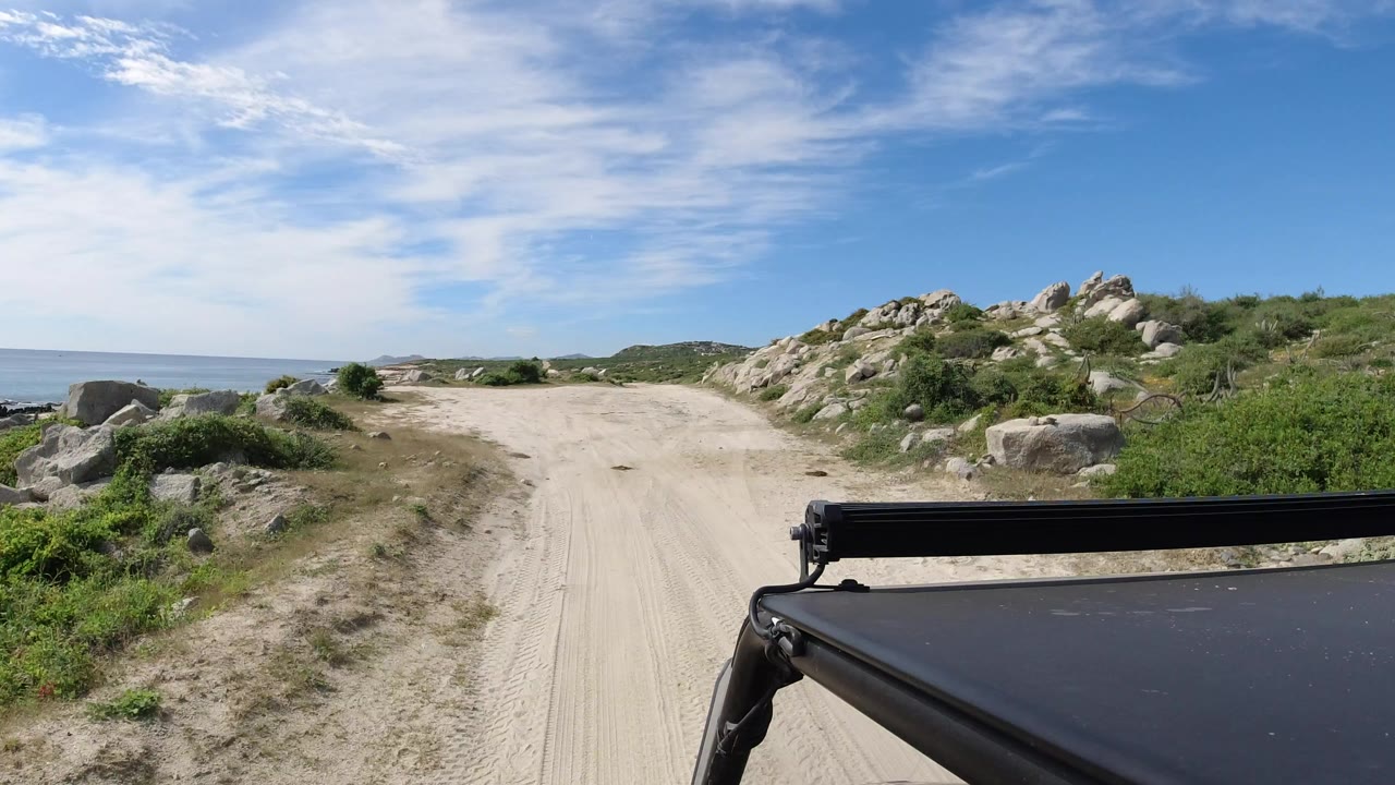 Los Cabos dirt road seen from a vehicle edge, with ocean visible on the left, rocky scrubland on the right, bright sky above, and no visible people.