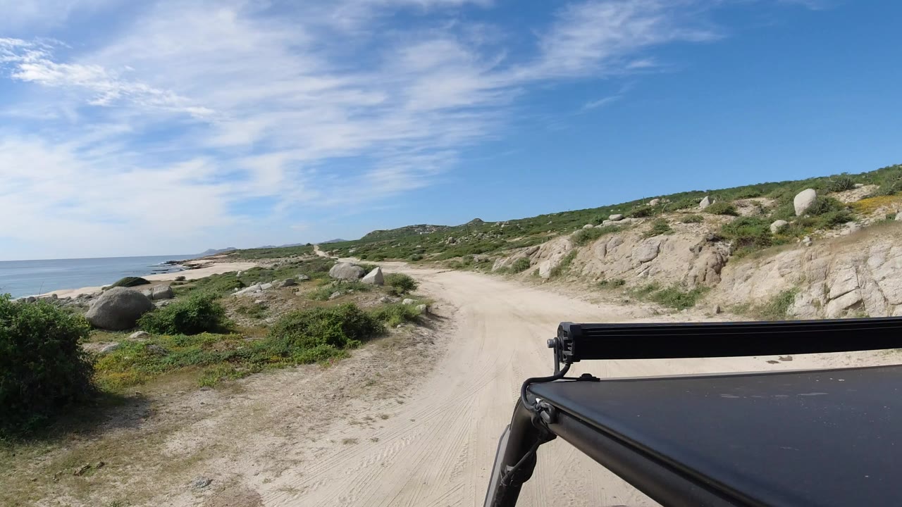 Start of a Los Cabos, Mexico vehicle POV showing a sandy road curving past pale rocks, green scrub, and ocean water on the left, with no visible people.