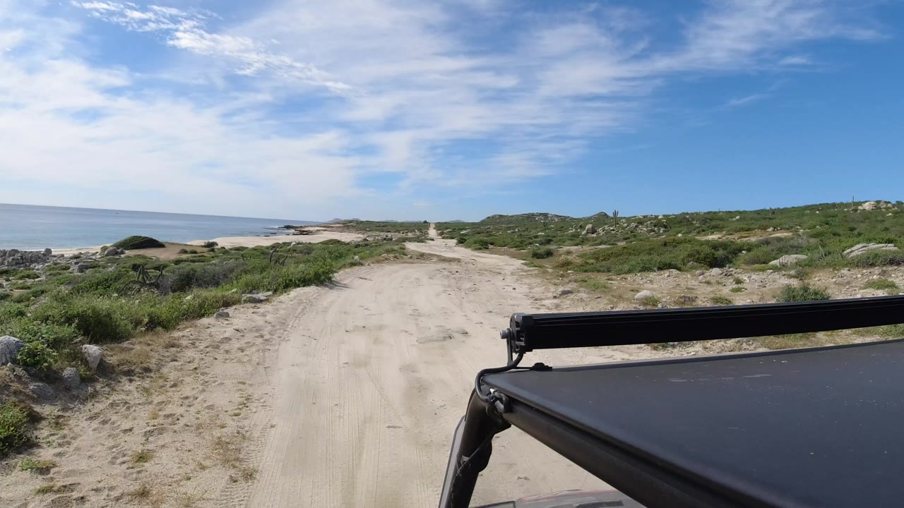 Start of a Los Cabos vehicle-perspective shot on a sandy road beside low scrub and the ocean, with no visible people.
