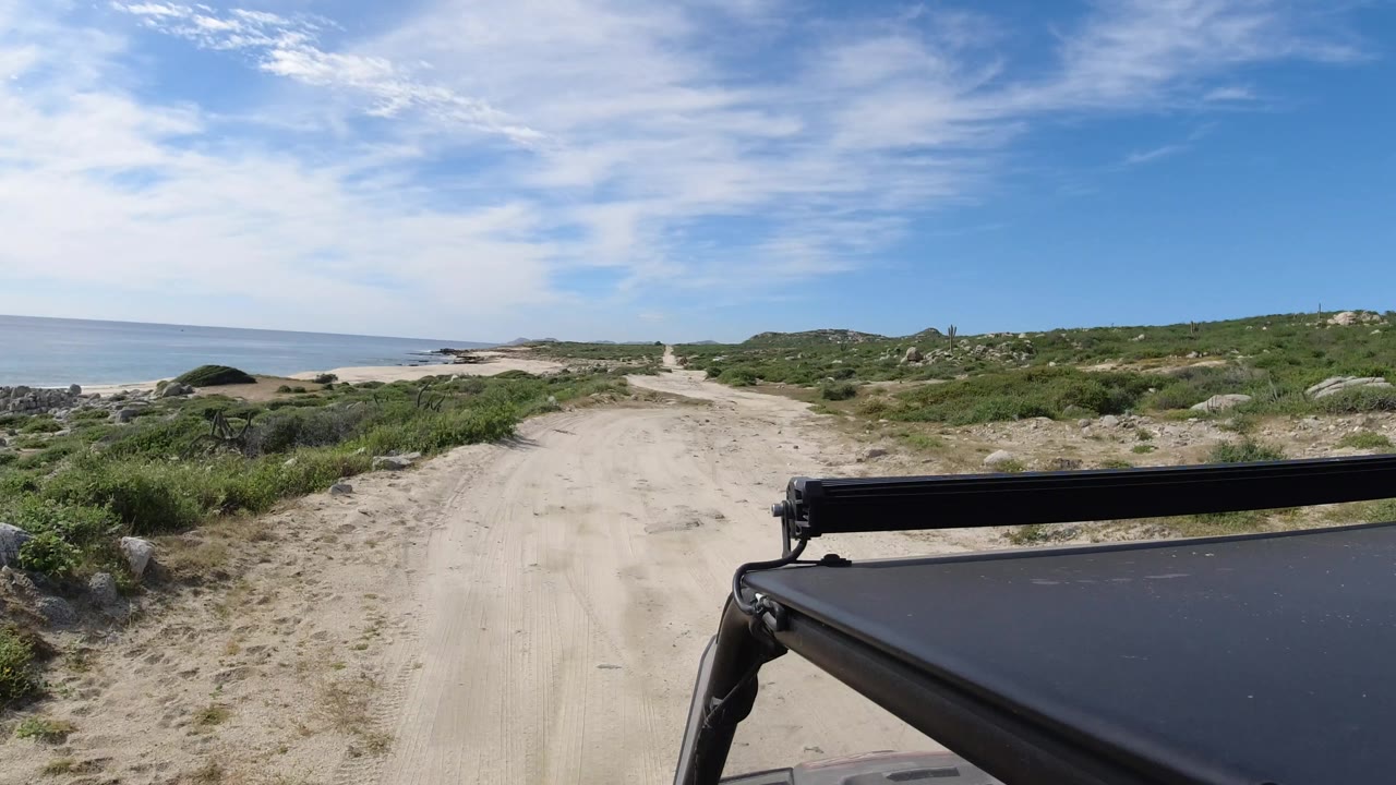 A sandy Los Cabos road continues ahead with ocean water on the left, scrubby dunes on both sides, and no visible people.
