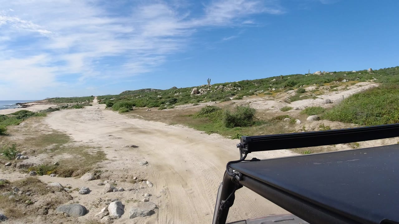 A sandy off-road track in Los Cabos, Mexico runs past low green brush toward the sea, with a black vehicle roof or light bar at the lower right and no visible people.