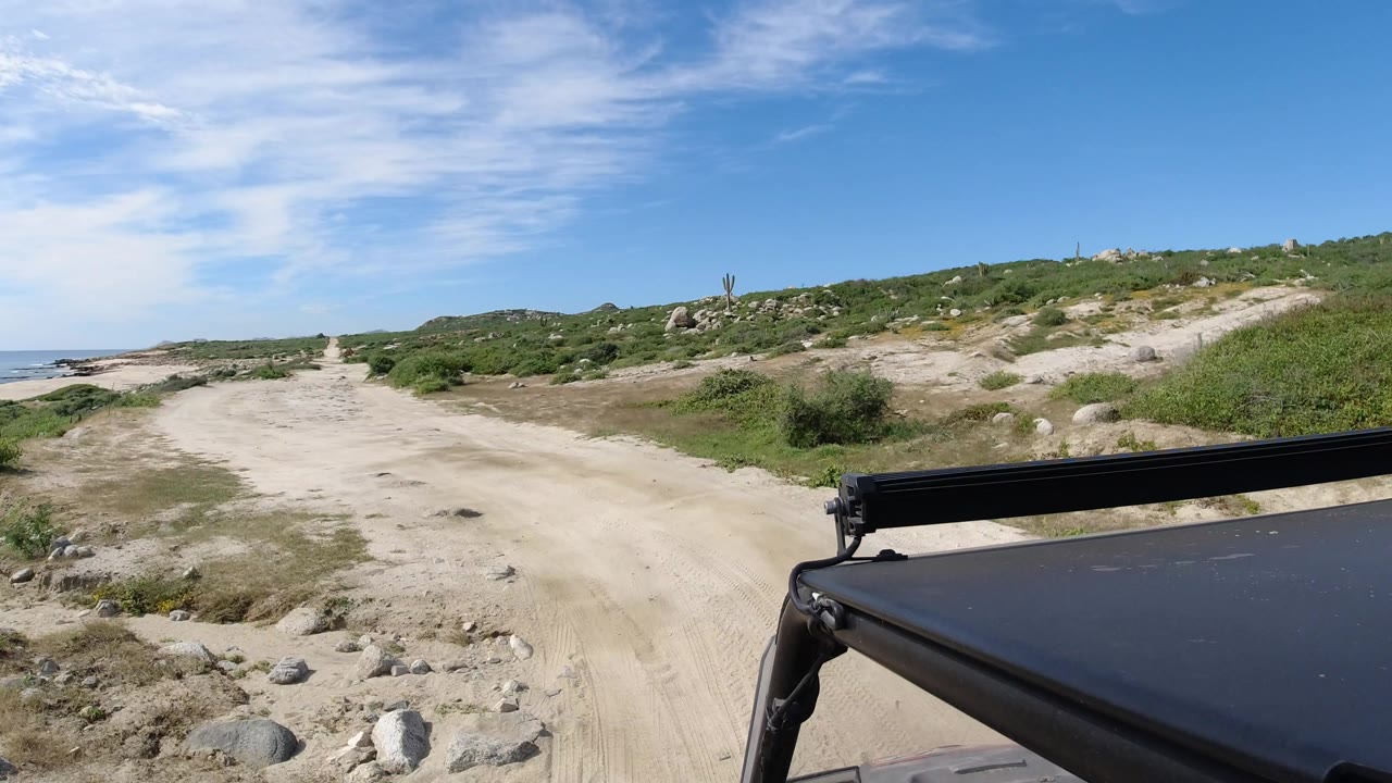 The vehicle viewpoint continues along the sandy Los Cabos track, with low brush on both sides, a pale sea edge at left, and no visible people.