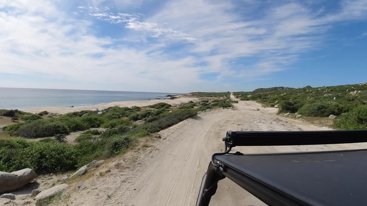 The vehicle foreground remains steady as the sandy track curves through low coastal vegetation toward rocky beach and ocean in Los Cabos, Mexico, with no visible people.