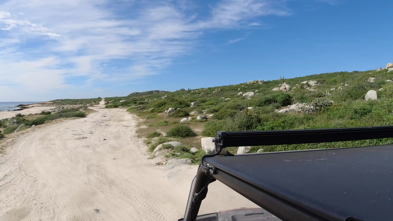 Start of a Los Cabos coastal drive showing a sandy road, low green scrub, scattered boulders, ocean at left, blue sky, vehicle roof hardware, and no visible people.
