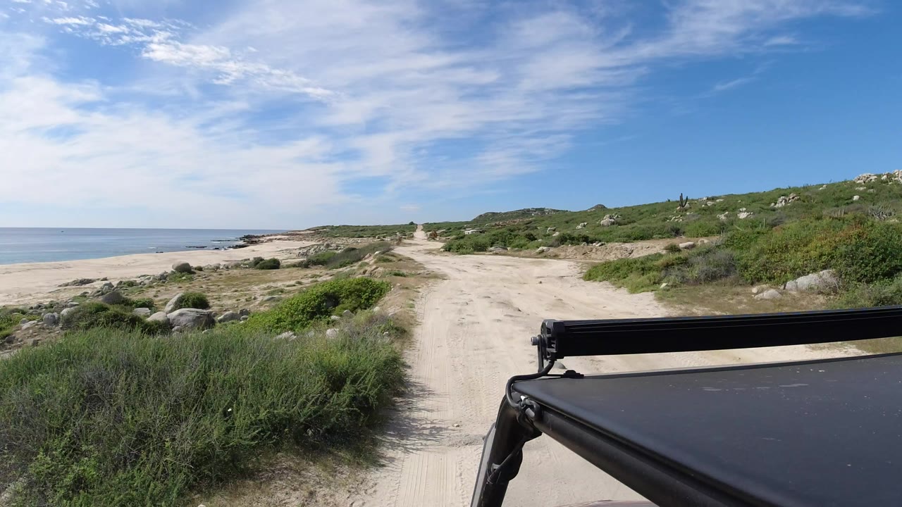 Forward view from a vehicle on a sandy Los Cabos beach track, with ocean on the left, green scrub on both sides, thin clouds overhead, and no visible people.