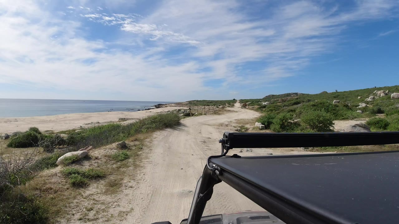 Opening frame of a sandy road beside the beach in Los Cabos, Mexico, with calm ocean at left, scrub-covered dunes at right, a dark vehicle roof in the foreground, and no visible people.