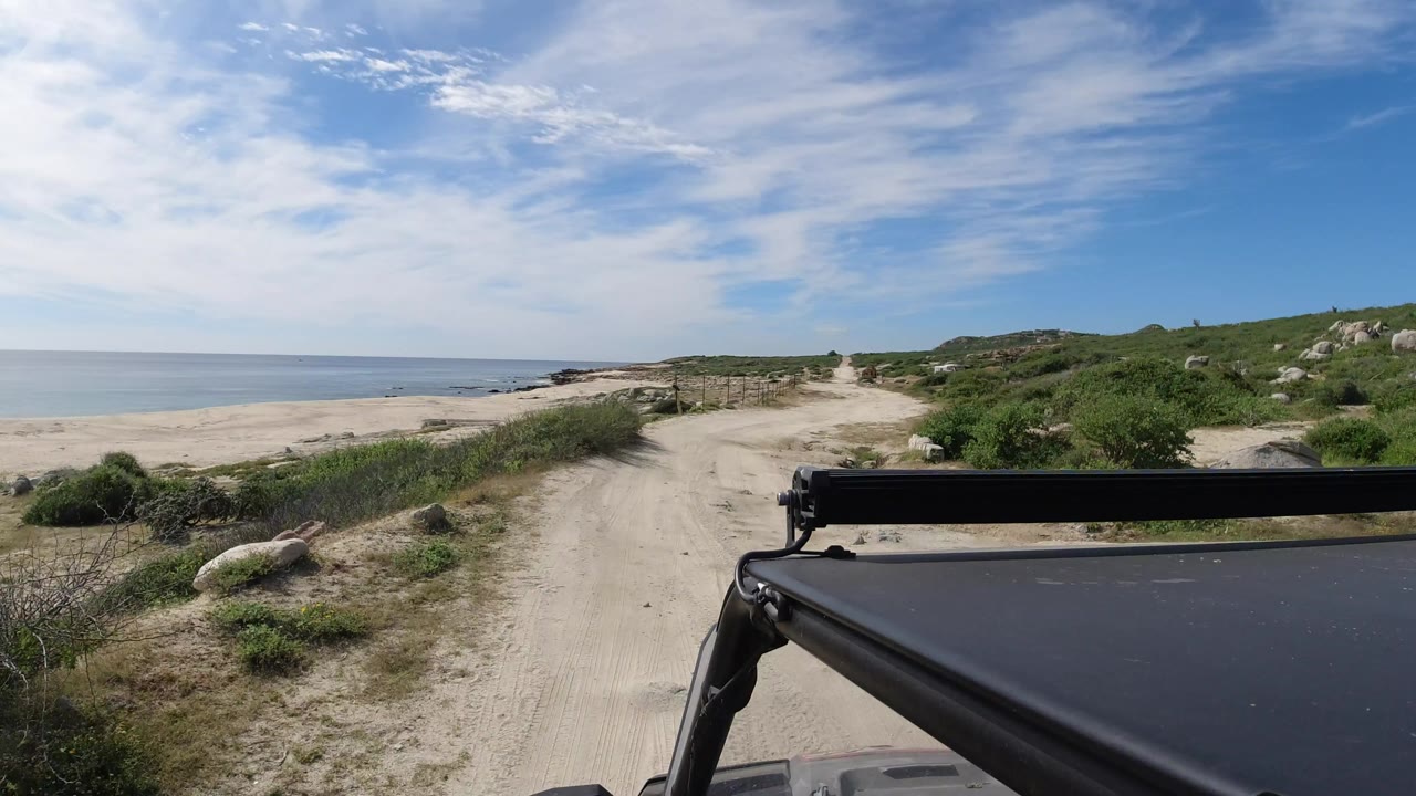 The vehicle POV continues down the pale sand road in Los Cabos, Mexico, with the beach and water opening to the left, green brush to the right, and no visible people.