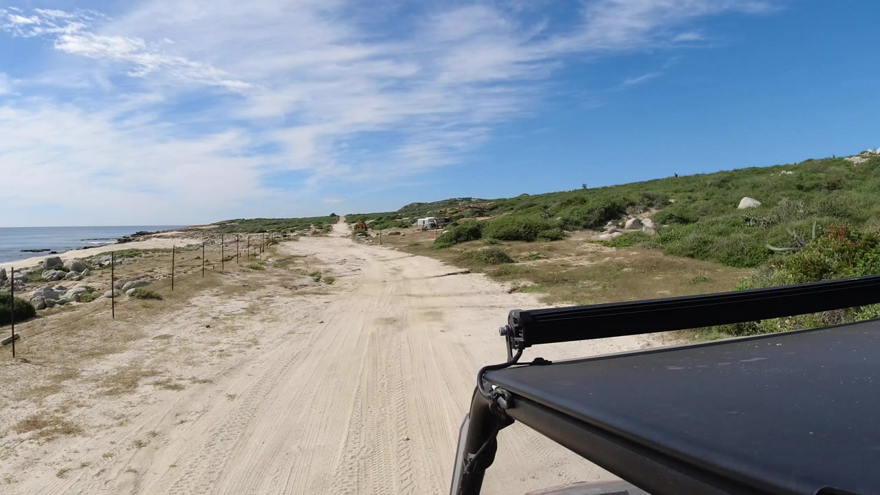 Sandy dirt road beside coastal water and green brush with a black vehicle roof in the foreground.