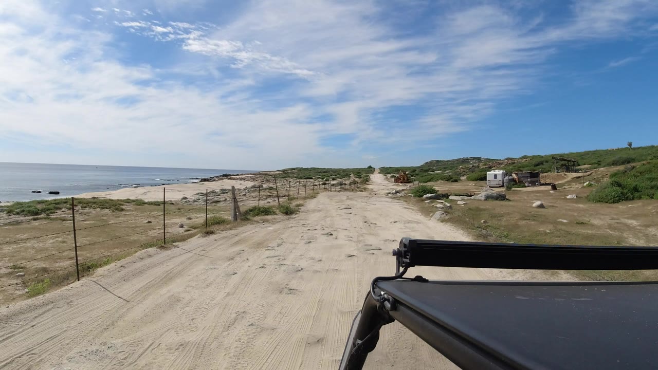 Start of a Los Cabos, Mexico dirt-road drive with the ocean on the left, fence posts along the sand, low green hills ahead, a dark vehicle edge in the foreground, and no visible people.