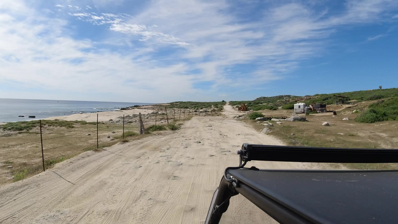 Los Cabos, Mexico coastal dirt track continues ahead under a blue sky, with the sea to the left, scrub-covered hills to the right, a dark vehicle edge at bottom right, and no visible people.