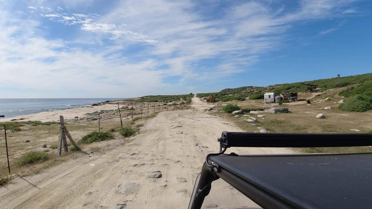 Coastal dirt road passes fence posts with ocean, shrubs, and small roadside structures.