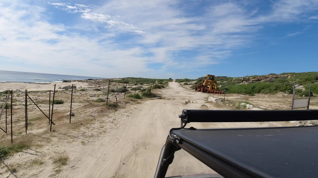Los Cabos sandy beach road with the ocean on the left, a yellow bulldozer farther ahead on the right side of the track, foreground Jeep hardware, and no visible people.