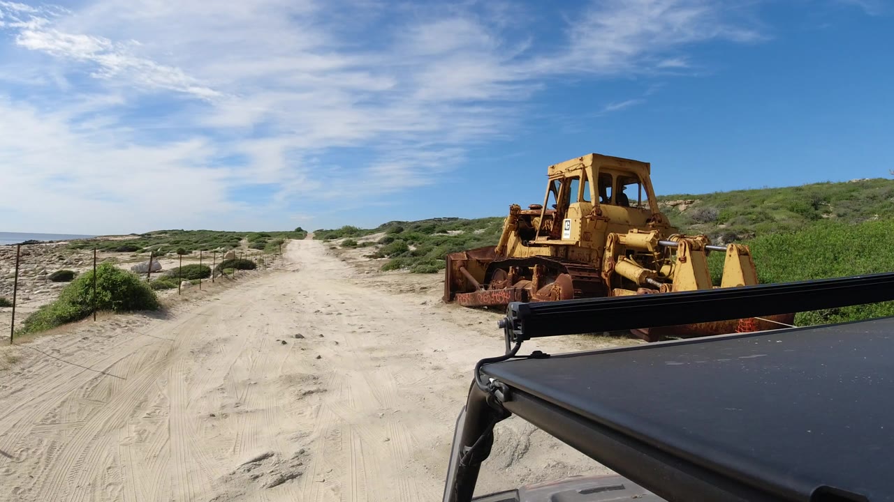 Sandy coastal road with yellow machinery beside a vehicle foreground.