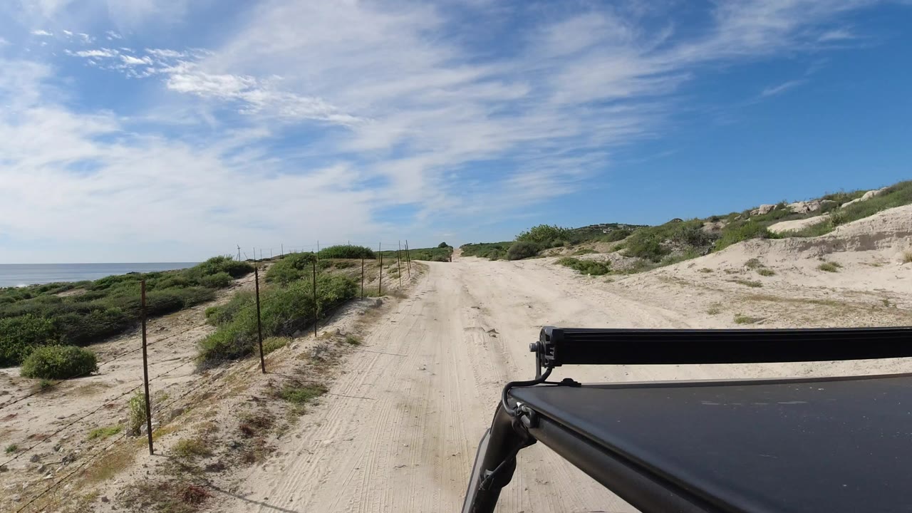 Opening view of a sandy road through low scrub in Los Cabos, Mexico, with the ocean on the left, a black vehicle edge at lower right, and no visible people.