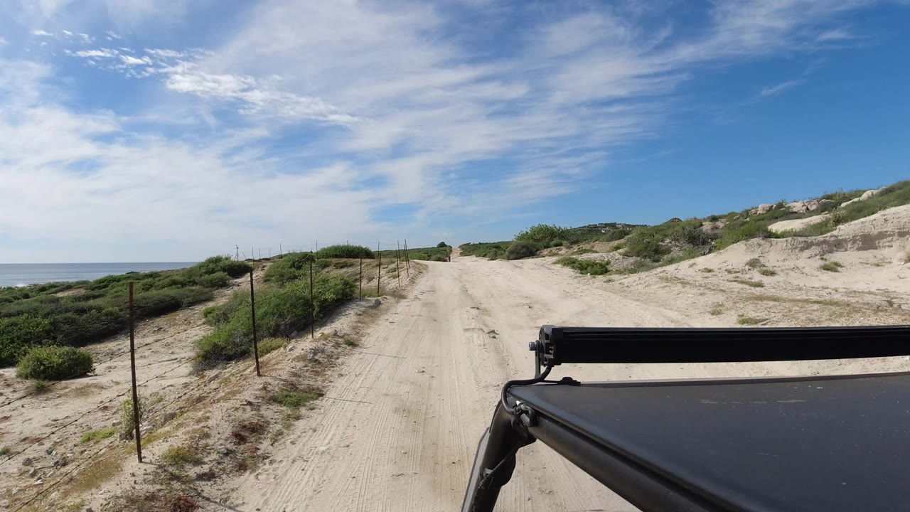 A similar Los Cabos road view shows the sandy track stretching ahead between scrub-covered dunes, with ocean still visible at left and no visible people.