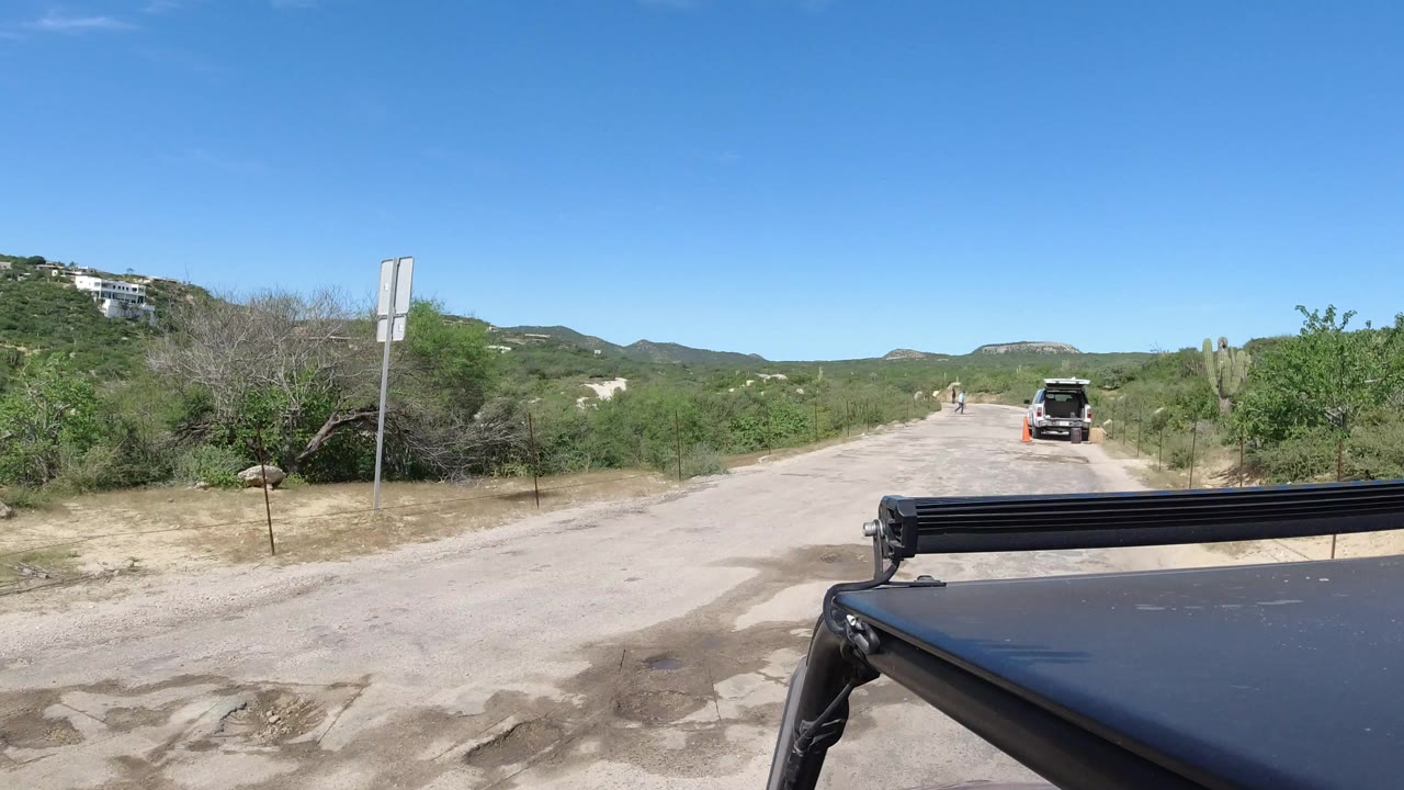 Dusty road with scrubland, a distant stopped vehicle, and a roof rack in the foreground.