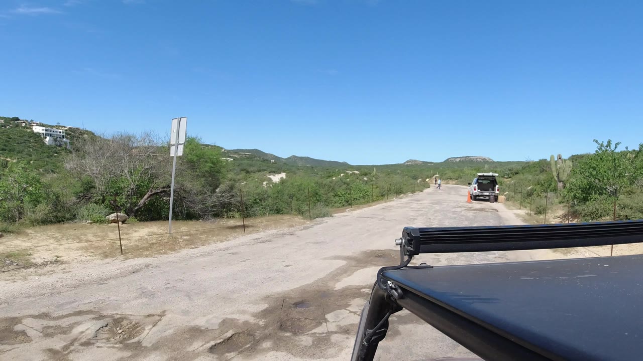 The Los Cabos road remains centered as the camera vehicle moves closer to a stopped SUV, with cactus and scrub along both sides and no identifiable people visible.