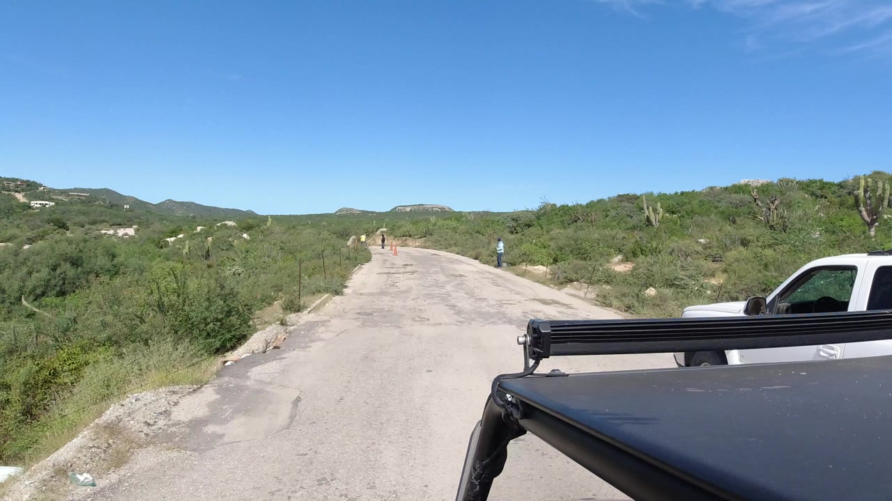 Vehicle roof beside a rough road through green scrub and low hills.