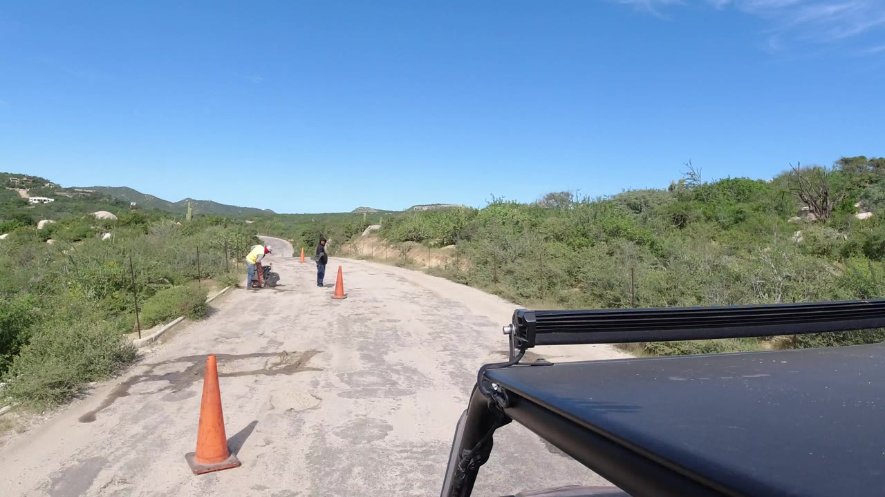 Forward view from a safari vehicle on a pale Los Cabos road, with workers and cones farther ahead, green scrub on both sides, and low hills under clear sky.