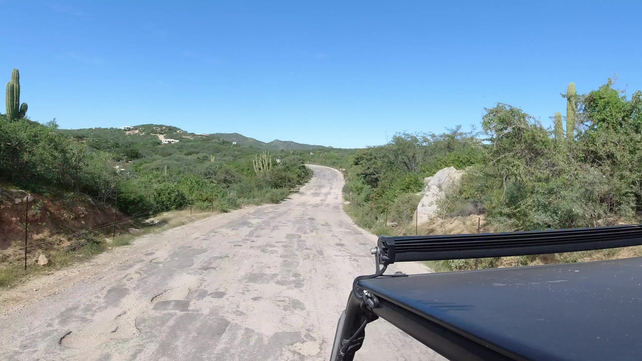 Los Cabos dirt road leading between green desert brush and cacti under a clear blue sky, with black Jeep hardware at the lower right and no visible people.