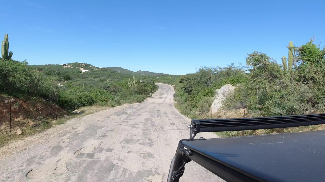 A pale, worn road continues through dense Los Cabos desert vegetation, with a Jeep roof edge and bar in the lower foreground and no visible people.