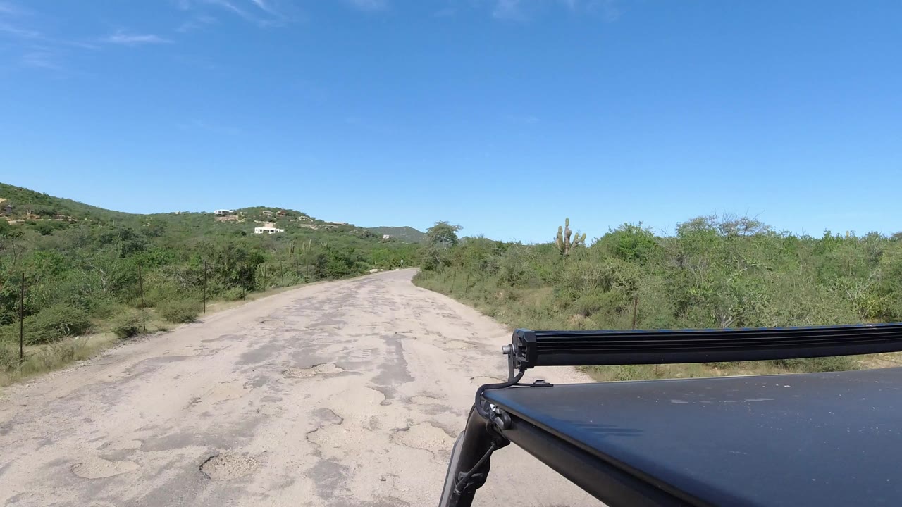 Cracked pale road through green scrub with vehicle roof gear in foreground.