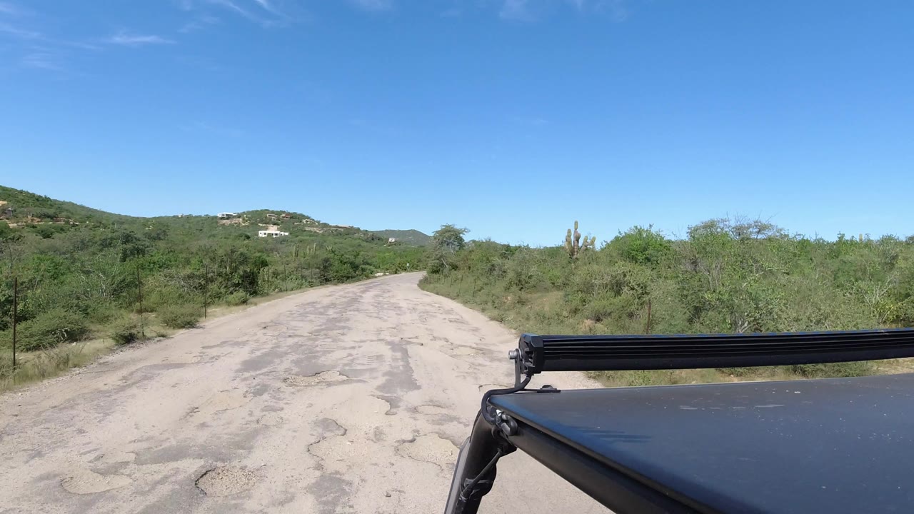 The road continues through Los Cabos scrubland, with the vehicle hood and black roof bar at the lower right, distant hills, and no visible people.