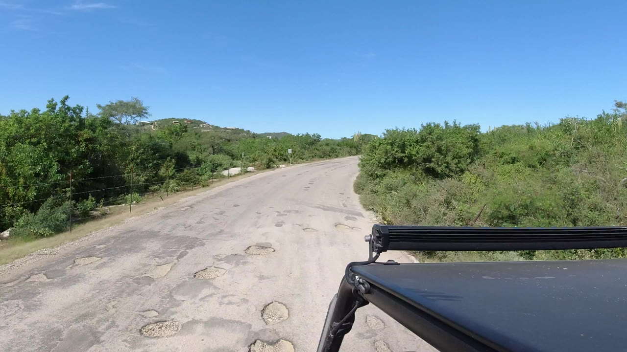 Forward-facing view from a vehicle on a sandy road in Los Cabos, Mexico, with patched pavement, green roadside brush, blue sky, and no visible people.