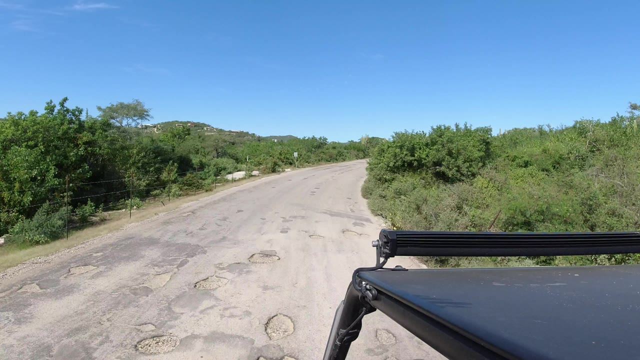 A slightly later view down the Los Cabos road shows the vehicle edge and roof bar at lower right, rough sandy pavement ahead, thick green brush, and no visible people.