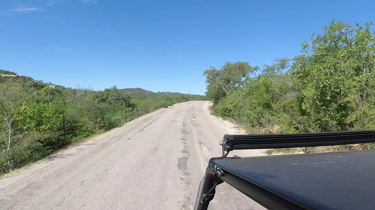 The road continues ahead in Los Cabos, Mexico, with dense shrubs on both sides, a black roof bar crossing the lower right, distant hills, and no visible people.