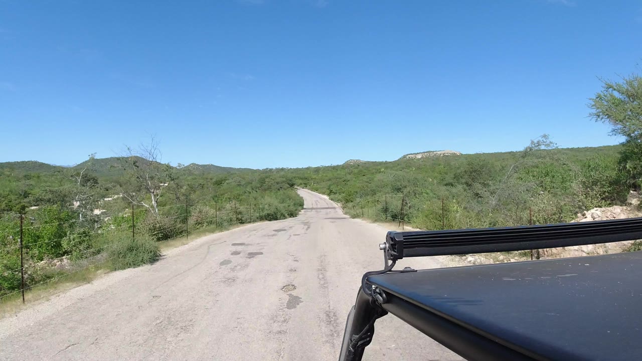 The road in Los Cabos continues through green brush with the horizon of low hills and a black roof rack visible across the lower right, with no visible people.