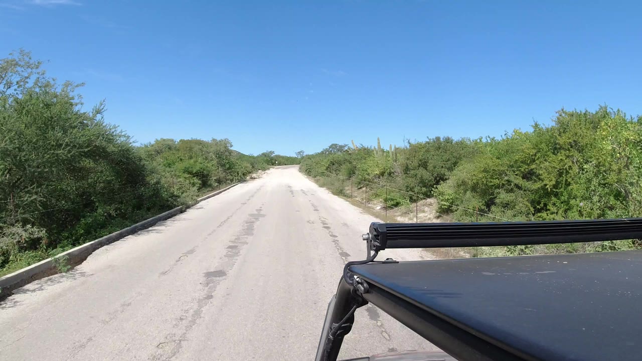 The Los Cabos road remains centered as dense brush lines both sides, distant low hills sit on the horizon, and the vehicle roof and light bar stay in the foreground with no visible people.
