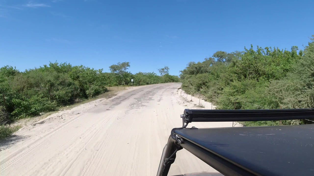 Forward vehicle view on a pale road in Los Cabos, Mexico, with green brush on both sides, blue sky above, and no visible people.