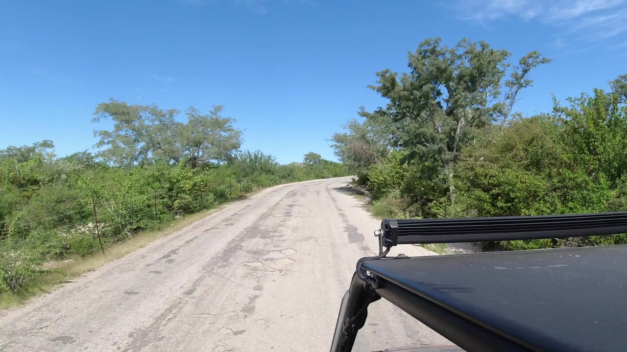 An empty pale road in Los Cabos, Mexico stretches ahead between green roadside brush, with a black vehicle roof or rack at the lower right and no visible people.