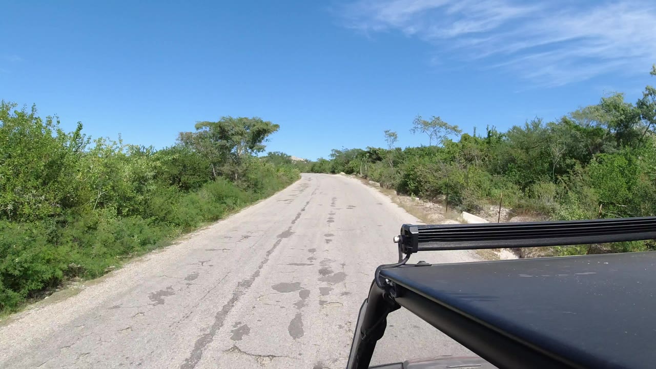 Vehicle-level view down a pale countryside road near Los Cabos, Mexico, with green brush on both sides, blue sky above, and no visible people.