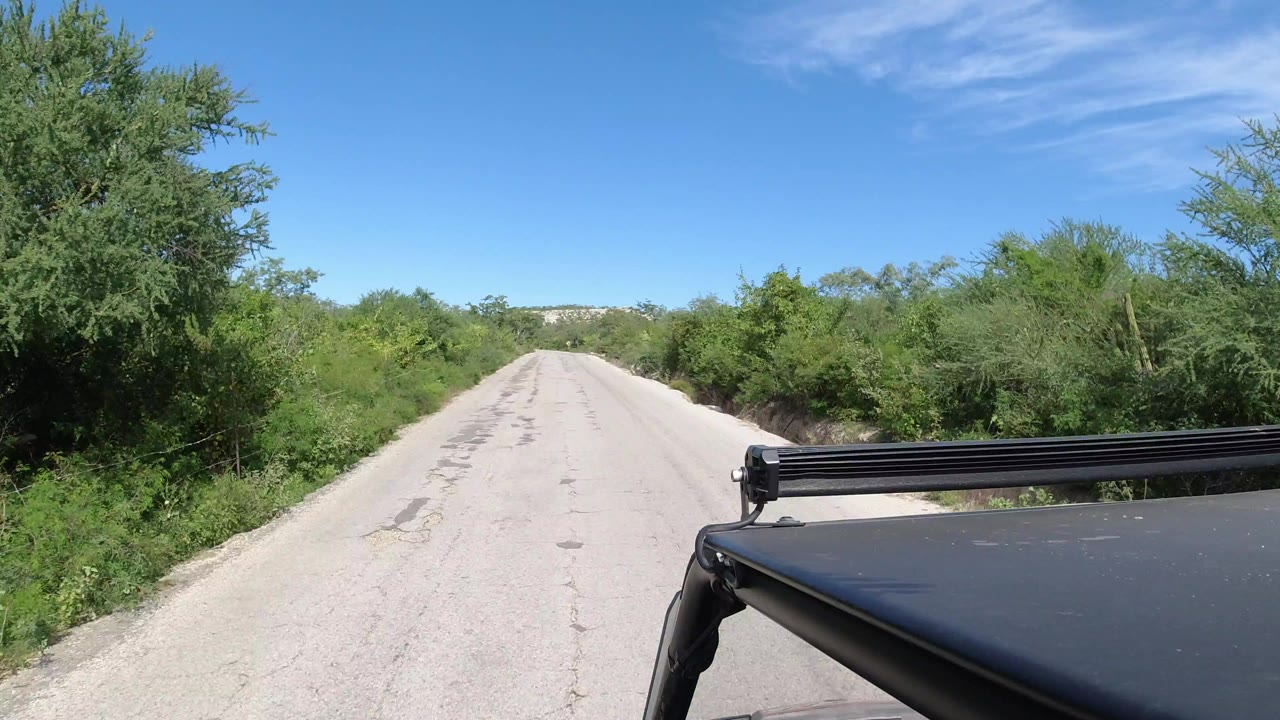 Opening view of a sandy road in Los Cabos, Mexico, bordered by green brush with the Jeep roof and light bar at lower right and no visible people.