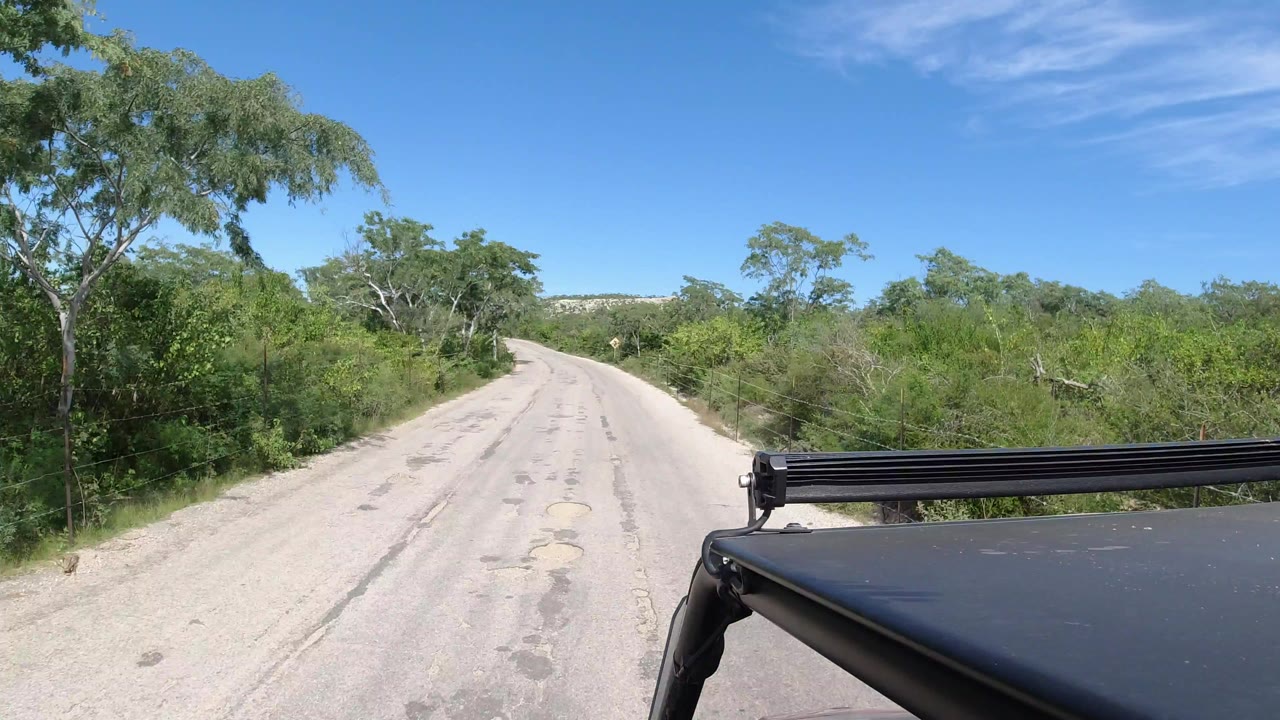 Start of a Los Cabos road view with an empty worn asphalt lane, green roadside trees, blue sky, and black vehicle hardware at lower right, with no visible people.