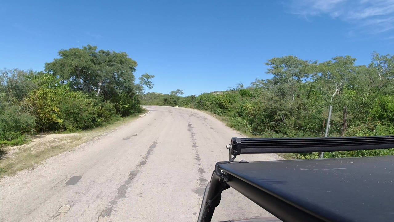 Opening frame of an empty Los Cabos road bordered by green trees, with a black vehicle roof rack across the lower right and no visible people.