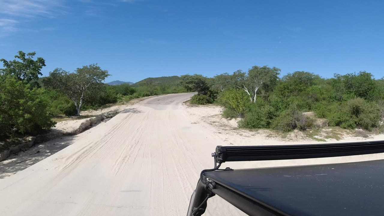Opening frame of a sandy Los Cabos road bordered by green trees, with distant hills, blue sky, a black vehicle edge in the lower right, and no visible people.