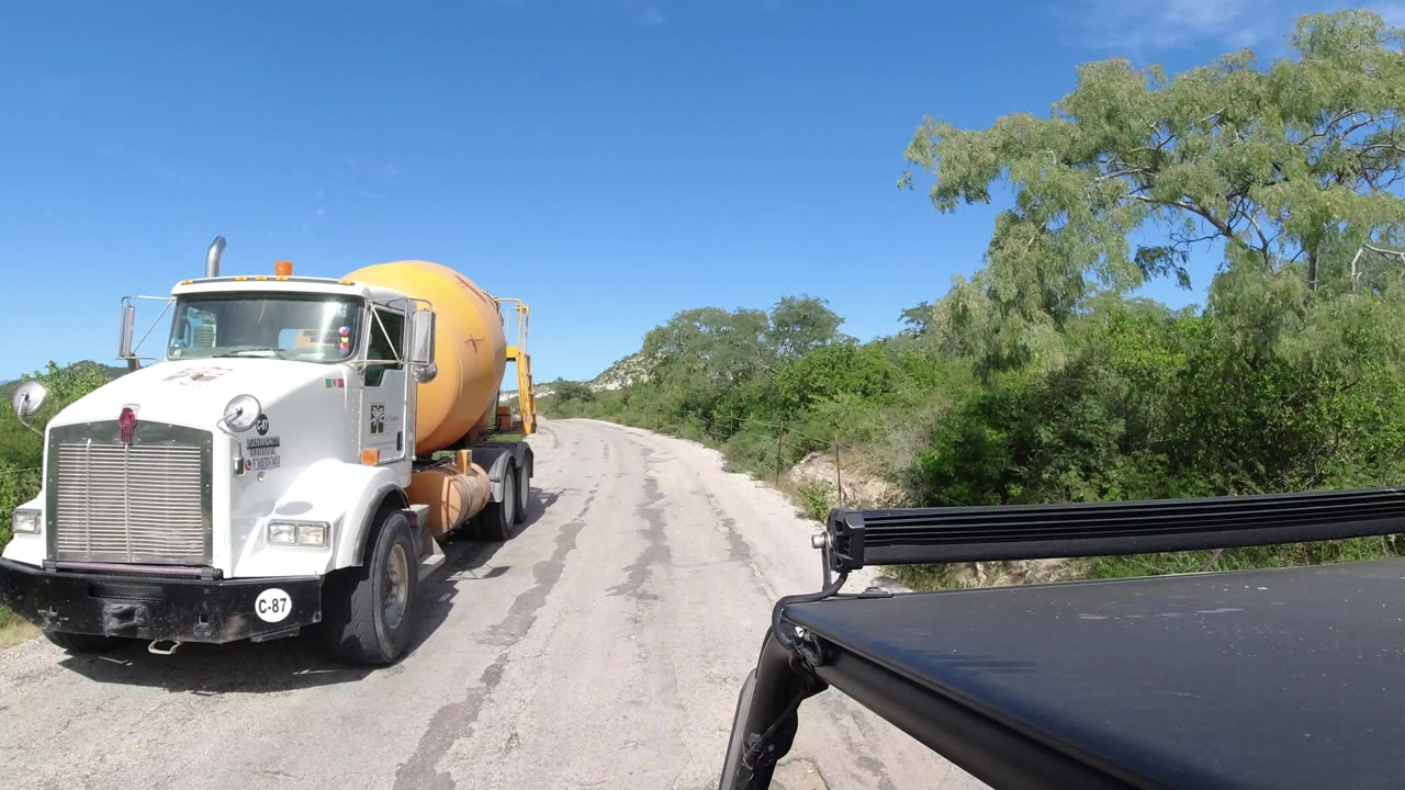 The cement mixer truck remains beside the camera on the Los Cabos road, with green trees, scrub, a clear blue sky, and no visible people in the frame.