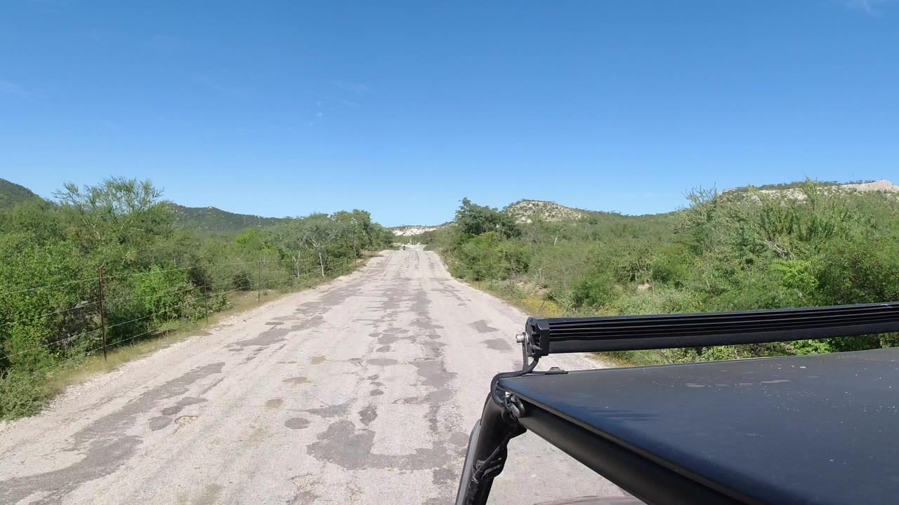 Los Cabos open road remains centered ahead, with a black roof edge at lower right, thick green roadside vegetation, distant rocky hills, and no visible people.