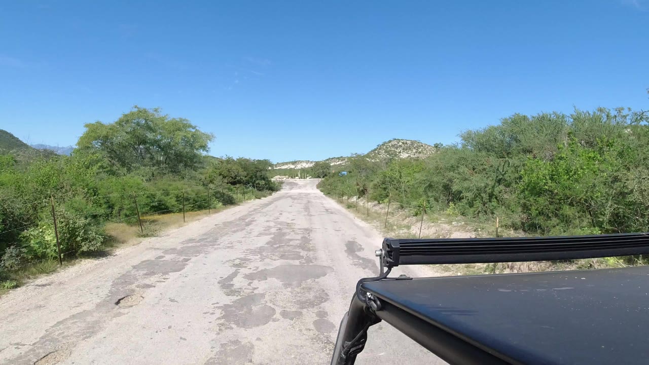 Opening frame of a Los Cabos, Mexico road seen from a vehicle, with dense green roadside trees, distant low hills, bright blue sky, and no visible people.