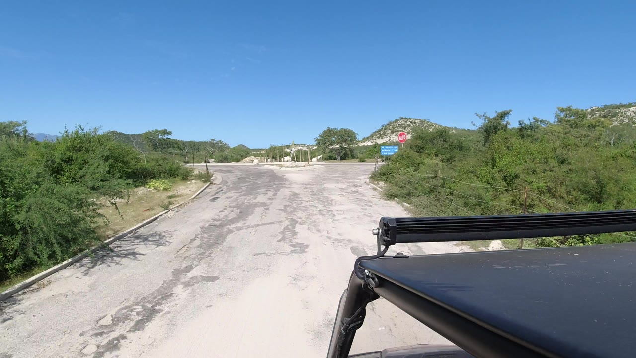 Los Cabos road approach with a red ALTO stop sign on the right, scrub vegetation around the curb, distant hills, and no visible people.