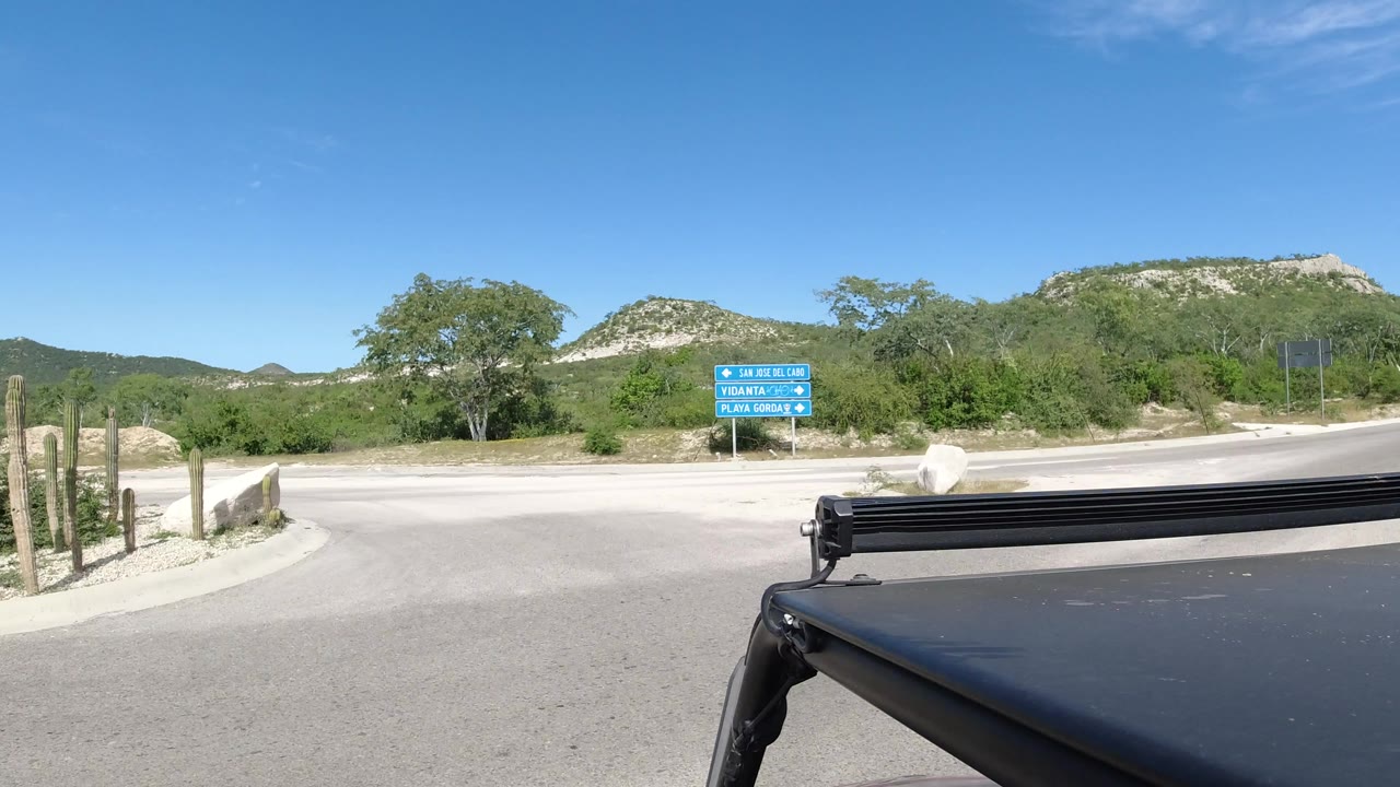 A slightly closer Los Cabos road view shows blue roadside signs ahead, pale pavement, brushy vegetation, low hills, and a fixed dark vehicle edge at the bottom, with no visible people.