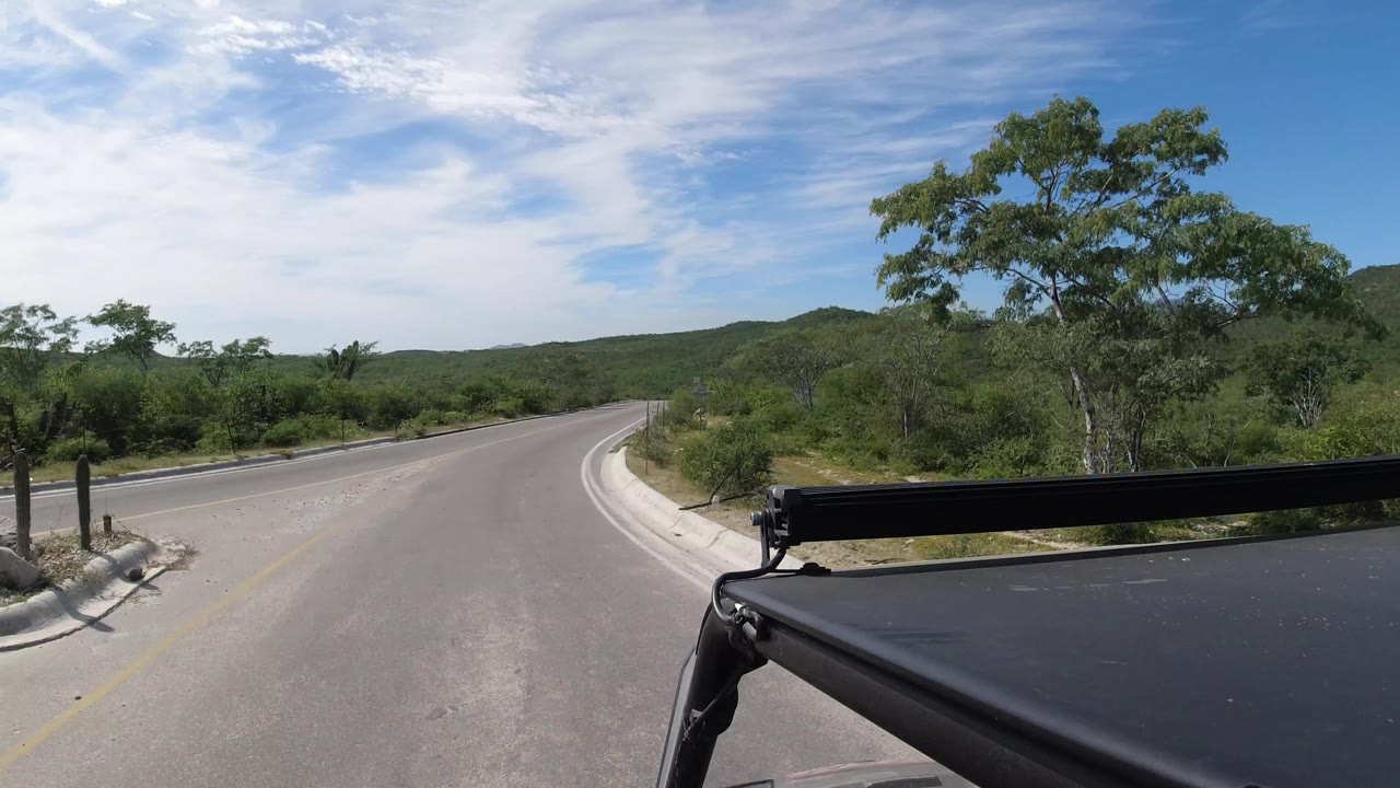 Empty curved road with green brush, low hills, and black vehicle hardware in the foreground.