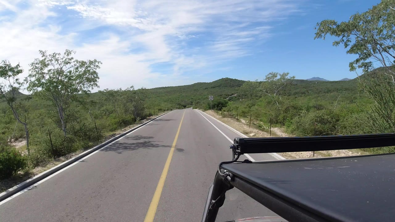Forward road view in Los Cabos, Mexico, showing a two-lane paved road, green roadside brush, low hills, blue sky, vehicle hardware at lower right, and no visible people.