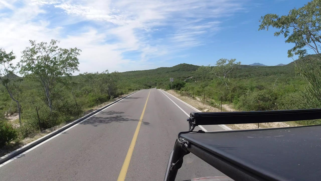 Los Cabos road view one second into the clip, with the center line leading toward low hills, dense green brush on both sides, a vehicle-mounted foreground edge, and no visible people.